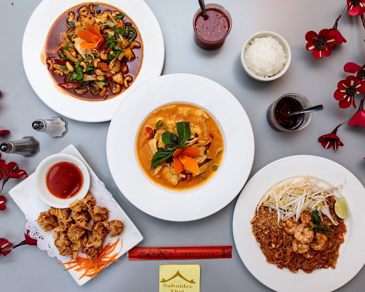 A table of Kob Jai Thai dishes: red curry, pad thai, crab rangoon, jasmine rice and sauces on white plates